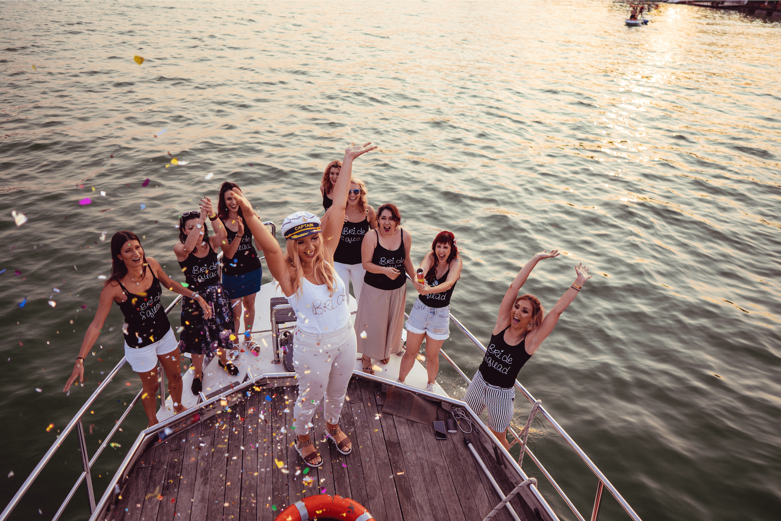 A group of women celebrate a bachelorette party on a boat at sunset. The bride, wearing a white outfit and captain’s hat, stands at the front throwing colorful confetti into the air while her friends in matching black “Bride Squad” tops cheer and raise their hands. The golden sunlight reflects off the water, creating a festive and joyful atmosphere.