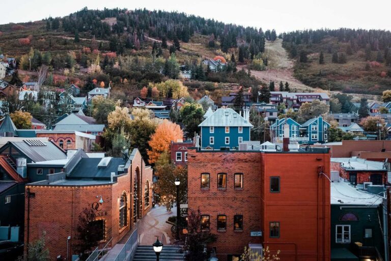 A charming view of Park City, Utah, with historic brick buildings in the foreground, colorful houses on the hillside, and a backdrop of autumn trees and forested mountains.