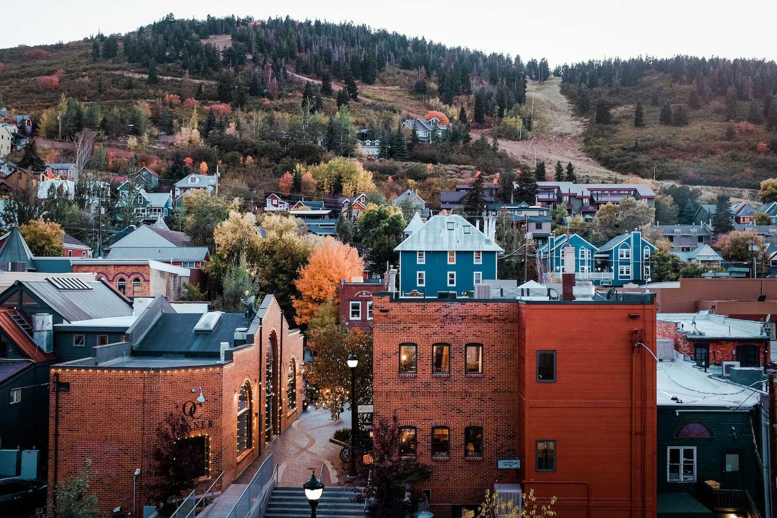 A charming view of Park City, Utah, with historic brick buildings in the foreground, colorful houses on the hillside, and a backdrop of autumn trees and forested mountains.