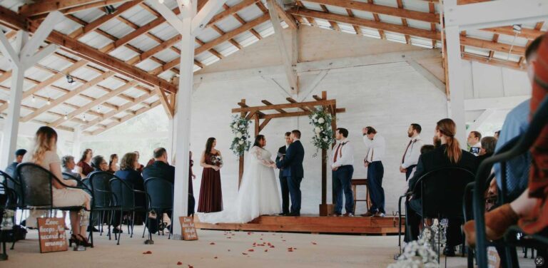 A bride and groom exchange vows beneath a wooden floral arch in a bright, rustic open-air barn venue.