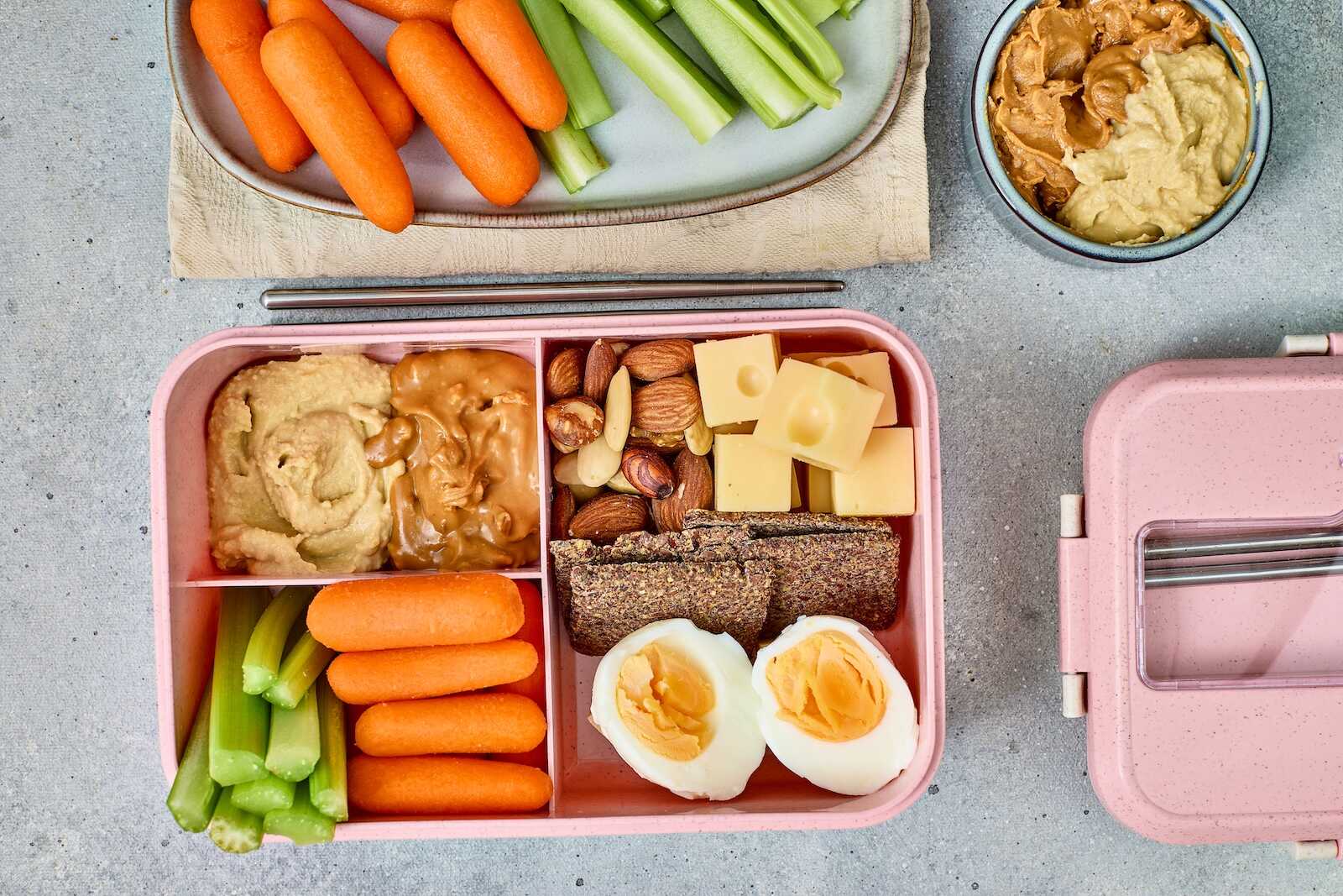 Two open bento boxes—one pink and one teal—each filled with cheese cubes, mixed nuts, crackers, baby carrots, celery sticks, hummus, and peanut butter, arranged on a gray surface. A small bowl of halved soft-boiled eggs sits to the right.